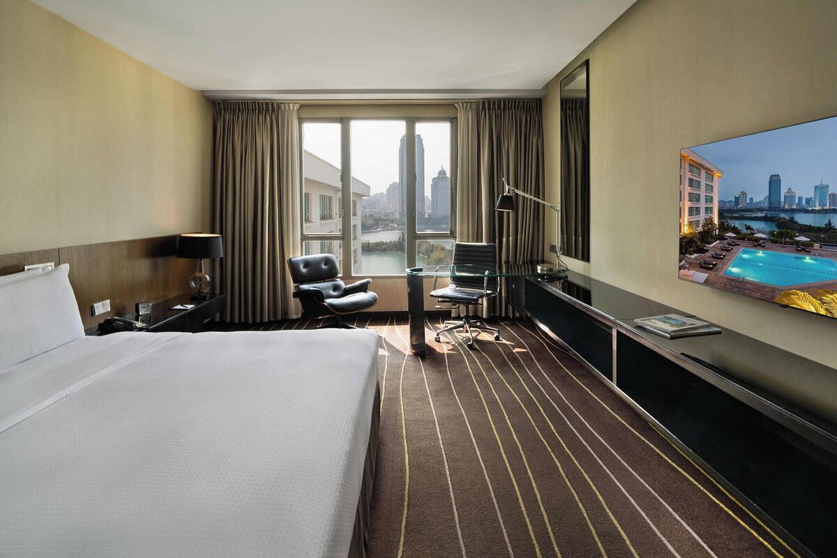 A hotel room with a large bed, a desk with a chair, a TV, and a view of a cityscape and pool.