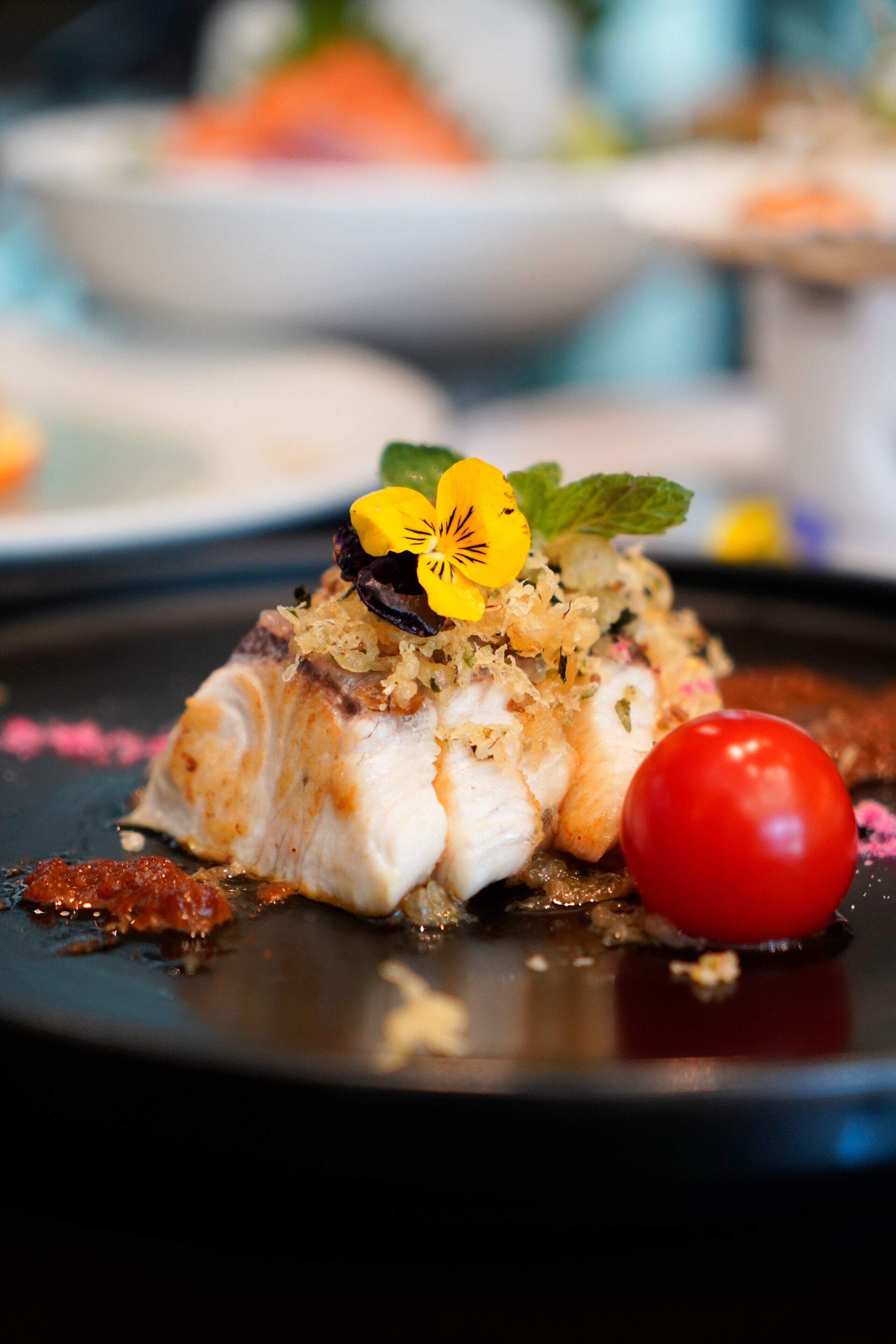 A dish with fish, garnished with flowers and a cherry tomato on a black plate.