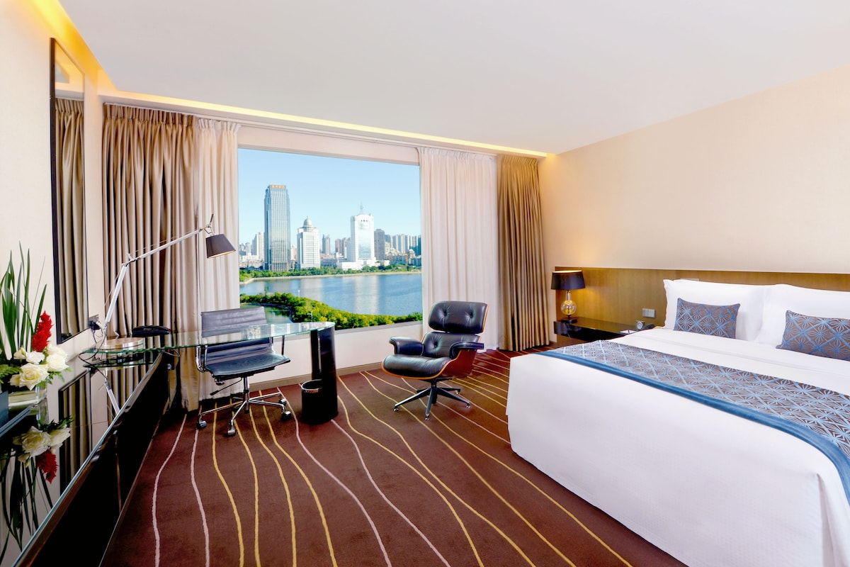 A hotel room with a large bed, a desk with a chair, a view of the cityscape, and a window with curtains.