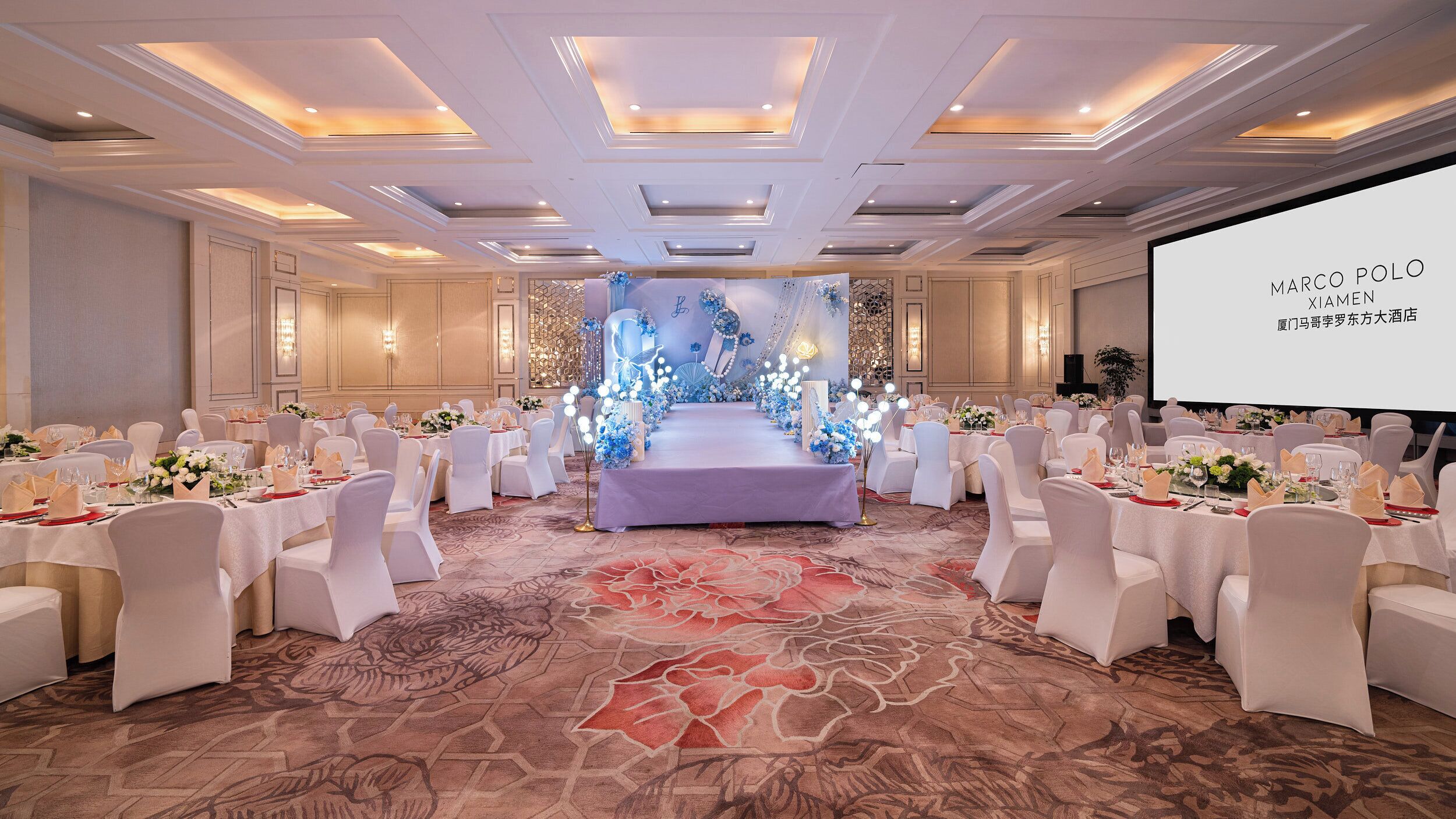A banquet hall with a central stage, white chairs, and tables set for a formal event.