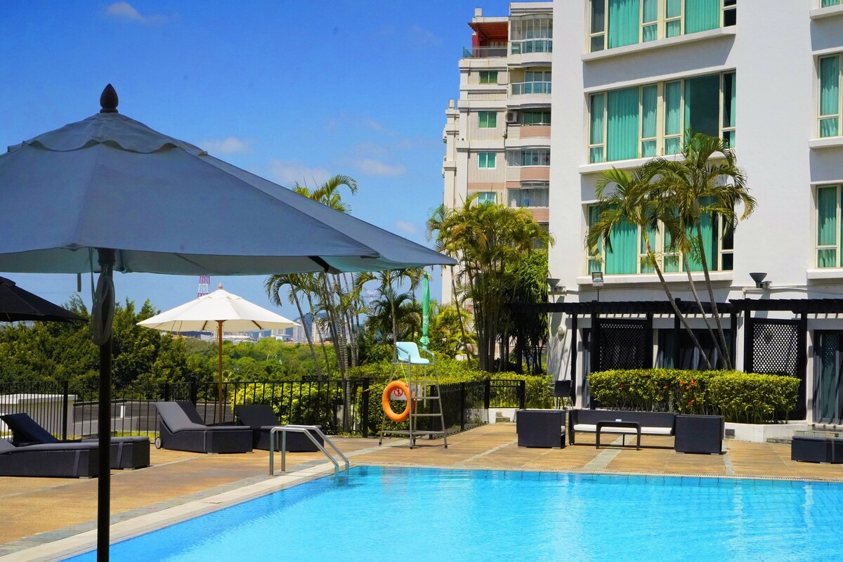 A swimming pool area with lounge chairs and umbrellas, surrounded by greenery and a multi-story building.