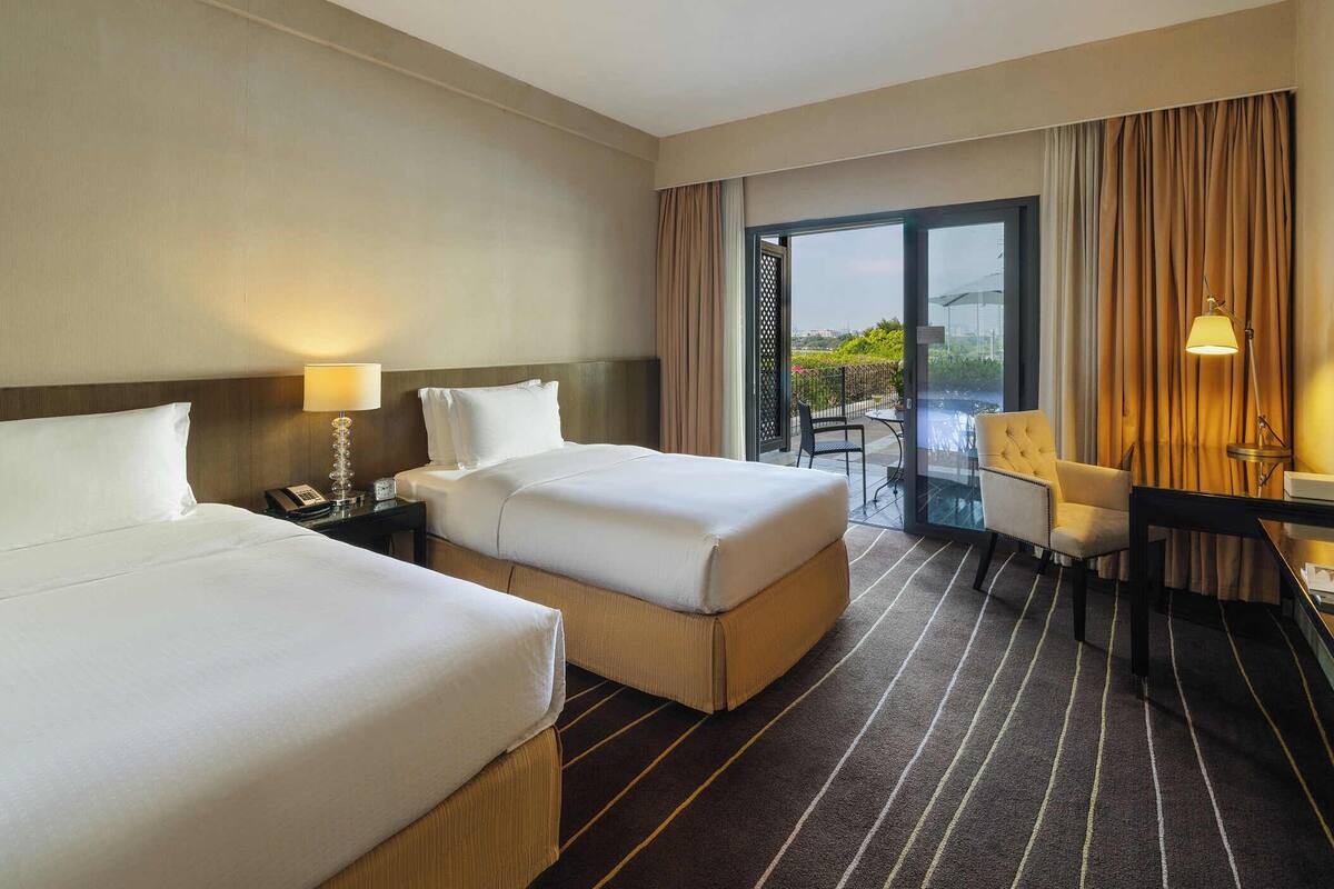 A hotel room with two beds, a desk, a chair, a phone, and a view of a cityscape and pool.