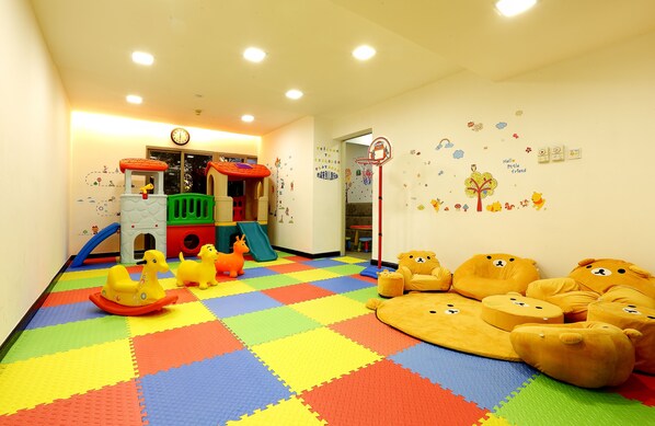 Uma sala de recreação infantil com tapetes de espuma coloridos, uma casinha e almofadas em formato de urso.