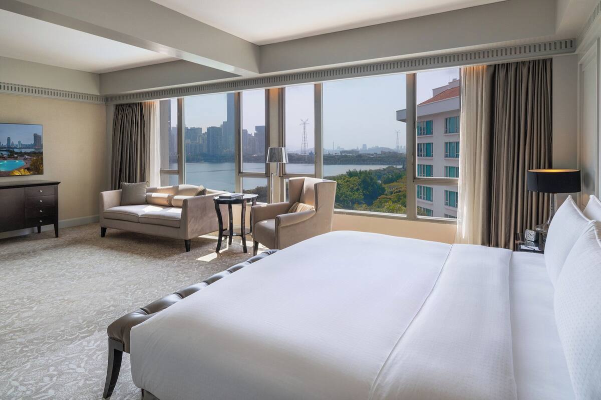 A hotel room with a large bed, two armchairs, a small table, and a view of a cityscape through large windows.