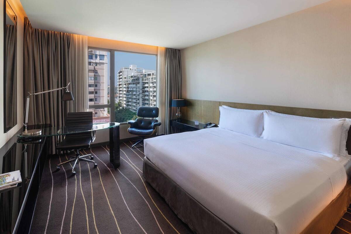 A hotel room with a large bed, a desk with a chair, a view of a cityscape, and a pool.