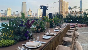 A rooftop dining area with a city skyline view, a long table set for a meal, and string lights.