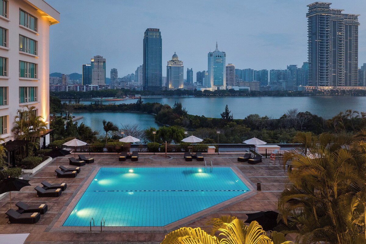 A rooftop pool with lounge chairs and umbrellas, overlooking a city skyline and a body of water.