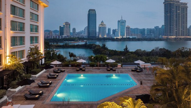 A rooftop pool with lounge chairs and umbrellas, overlooking a city skyline and a body of water.
