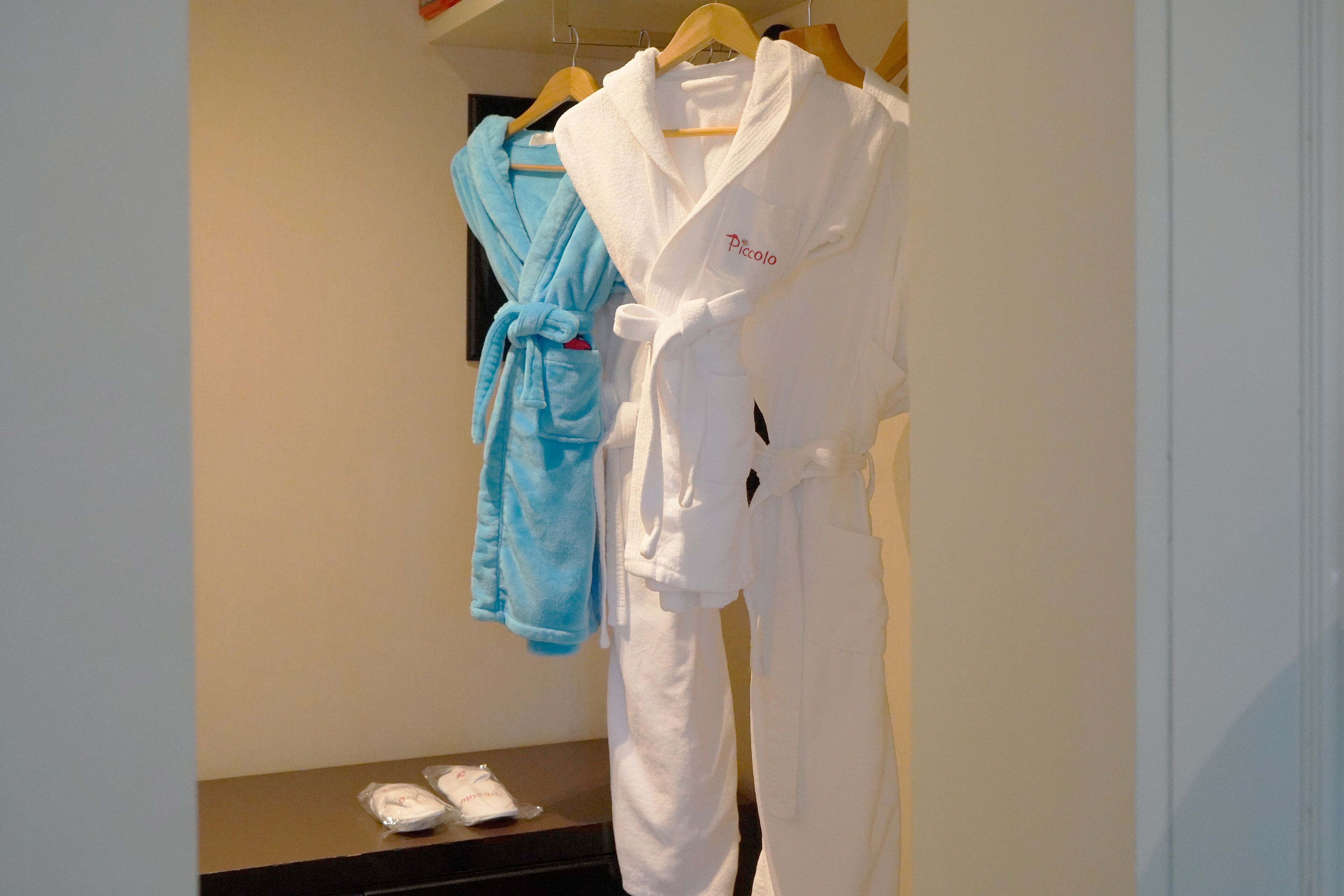 A hotel room with a bed, a small table with slippers, and two robes hanging on a rack.