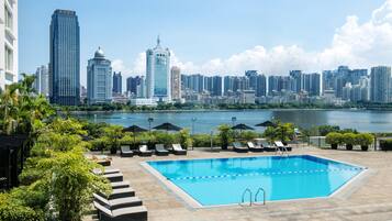 A swimming pool with lounge chairs and umbrellas, surrounded by greenery, with a city skyline in the background.
