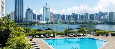 A swimming pool with lounge chairs and umbrellas, surrounded by greenery, with a city skyline in the background.