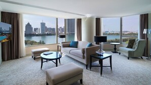 A spacious hotel room with a large window offering a city view, a sofa, a chair, a small table, and a lamp.