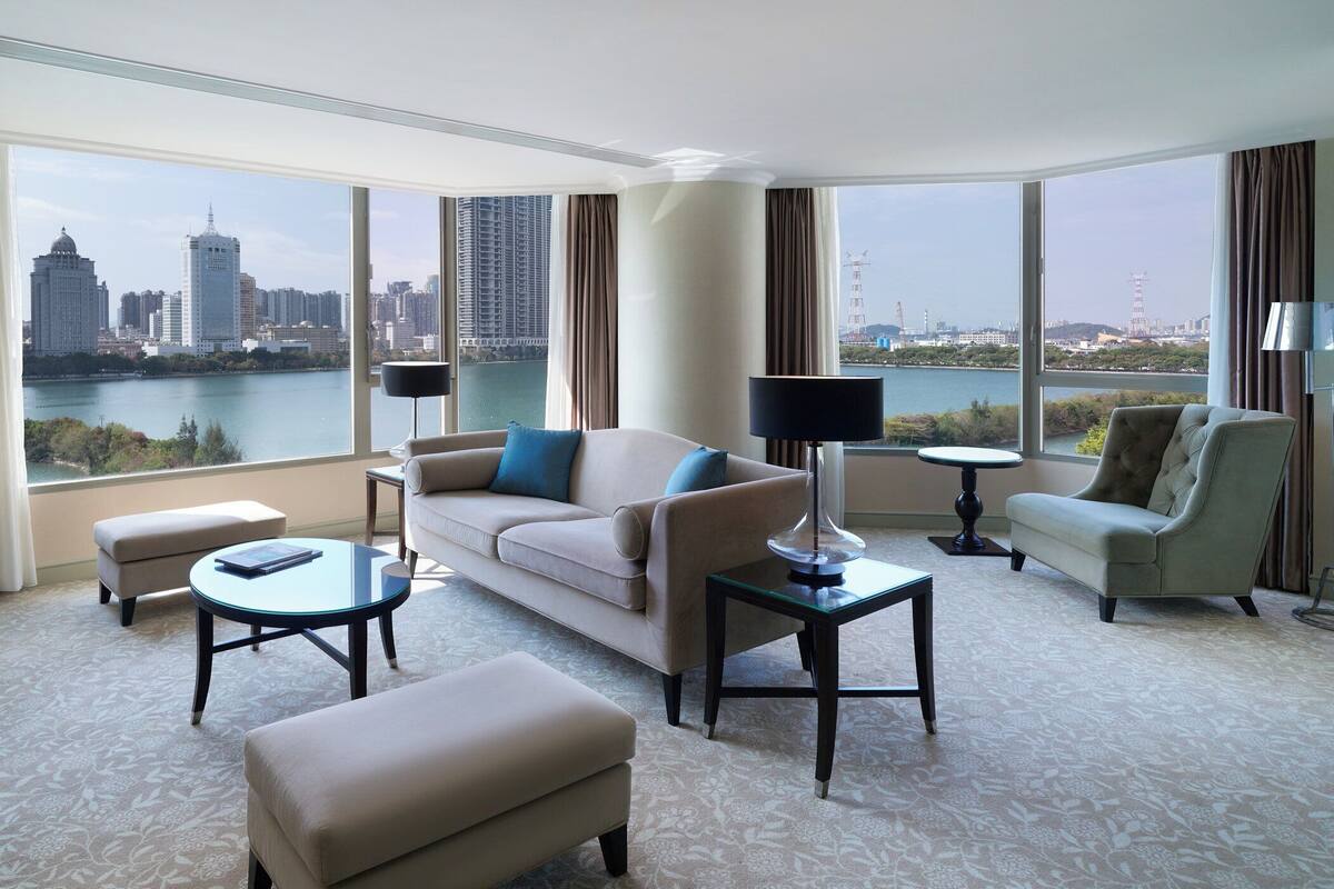 A spacious hotel room with a large window offering a city view, a sofa, a chair, a small table, and a lamp.