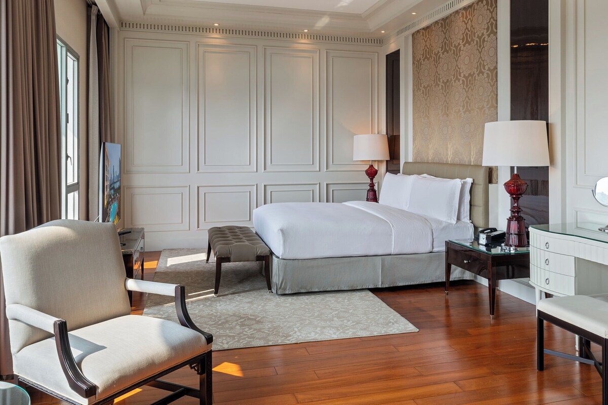 A hotel room with a large bed, two bedside tables with lamps, a chair, a desk with a mirror, and a large window with curtains.