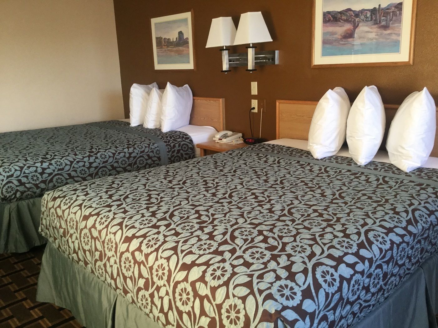 A hotel room with two beds, each with patterned bedspreads, a nightstand with a phone and lamp, and wall art depicting landscapes.