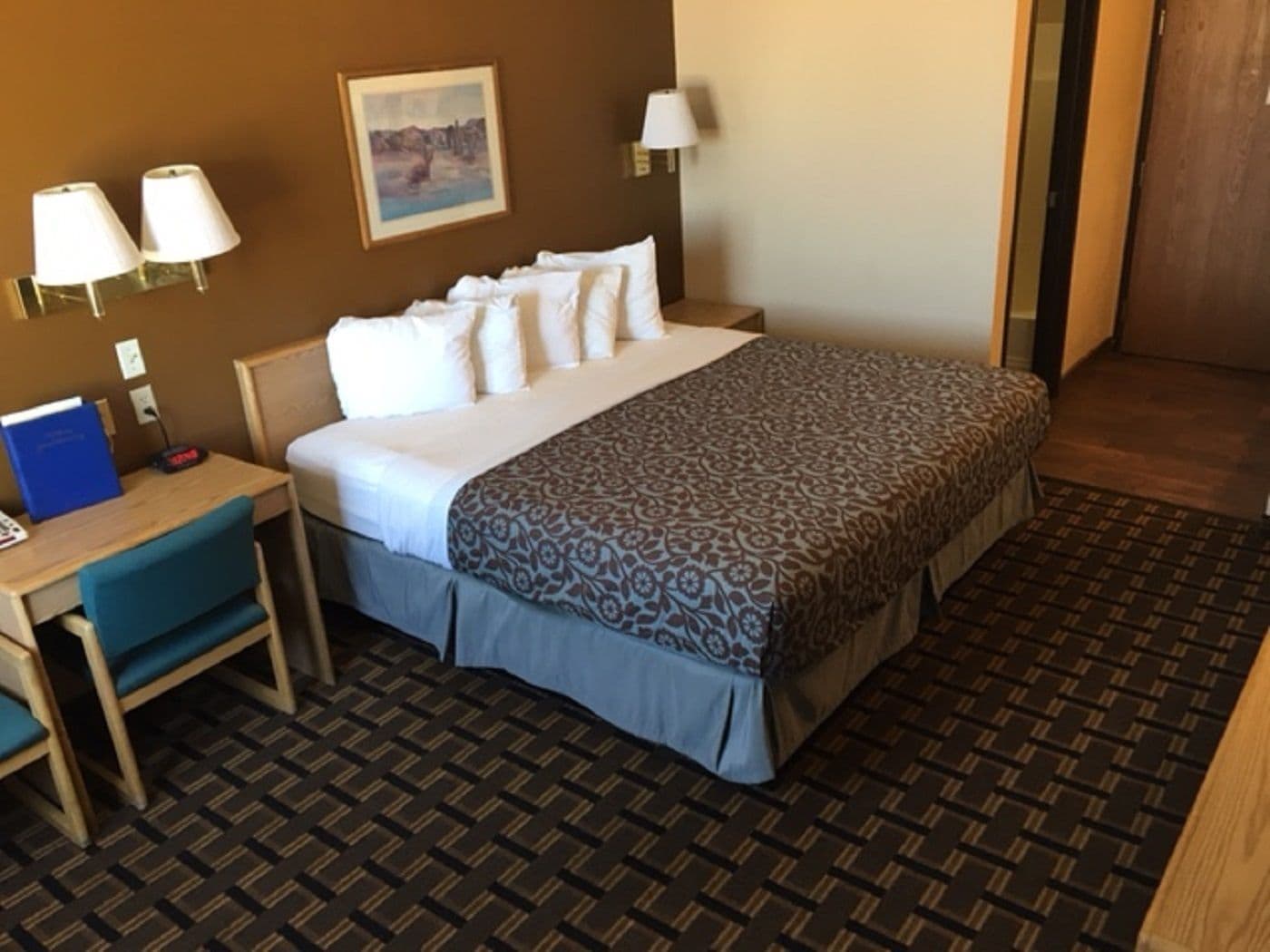 A hotel room with a bed, a desk, a chair, a lamp, and a painting on the wall.