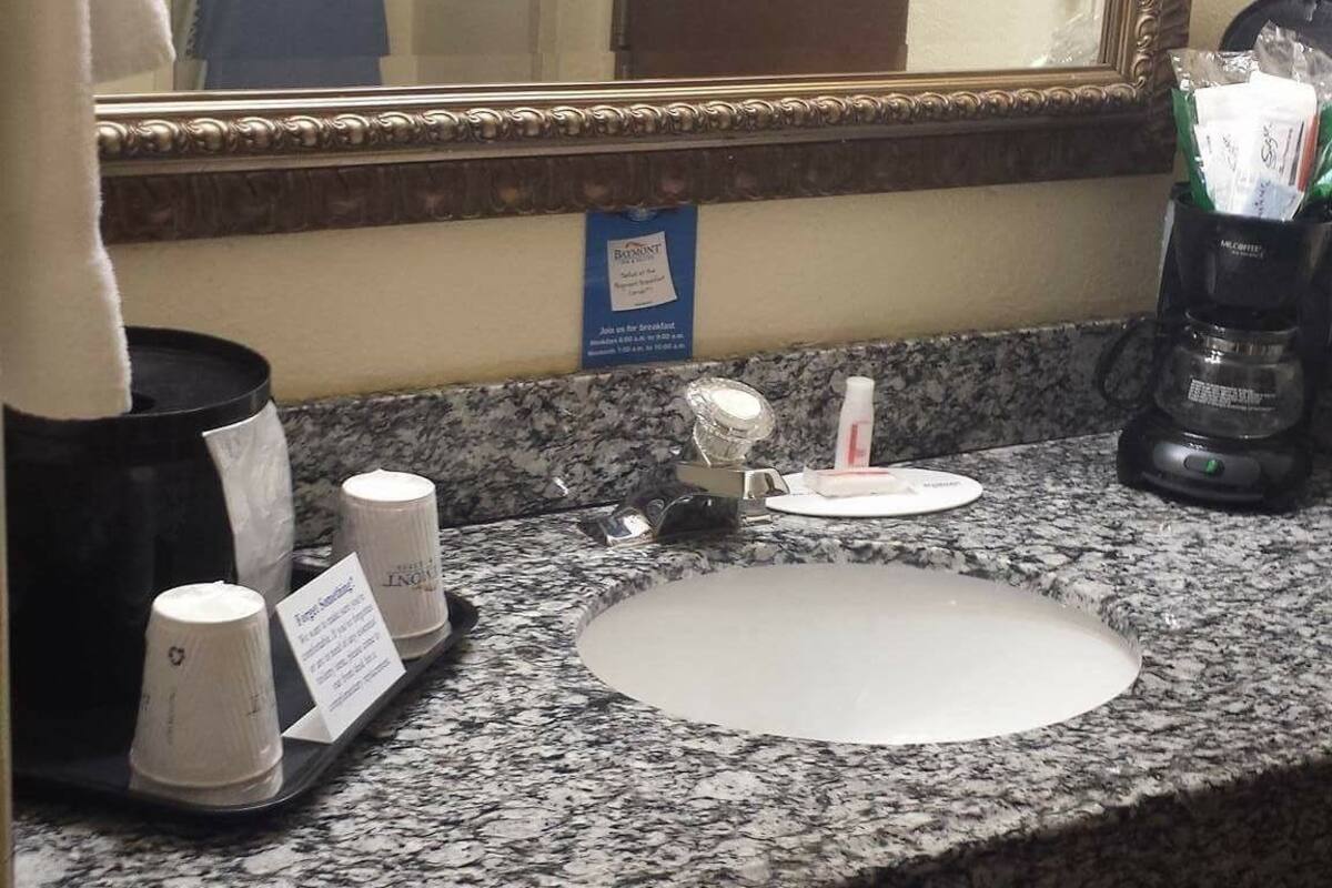 A hotel bathroom with a granite countertop, a sink, a mirror, a hairdryer, and a coffee maker.