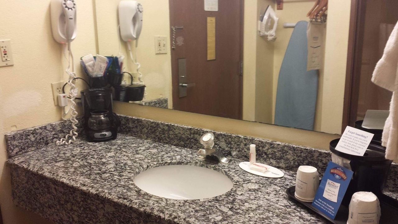 Casa de banho de hotel com bancada de lavatório em granito, secador de cabelo, espelho e máquina de café.