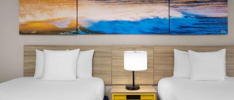 Habitación de hotel con dos camas, cabecero de madera, mesita de noche con lámpara y una gran pintura con vista al océano.