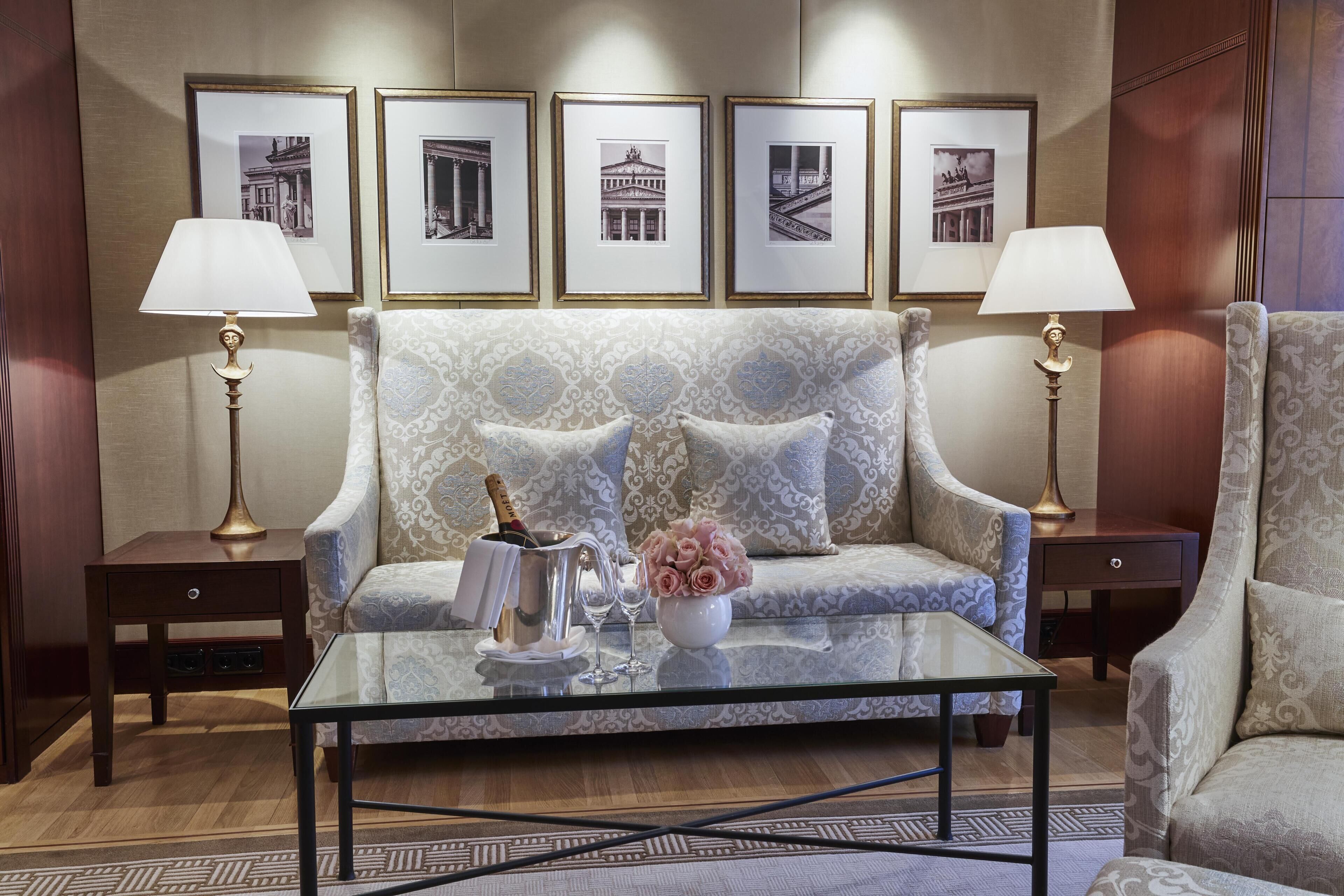 A room with a sofa, two lamps, a glass coffee table with a bottle and glasses, and a vase with flowers.