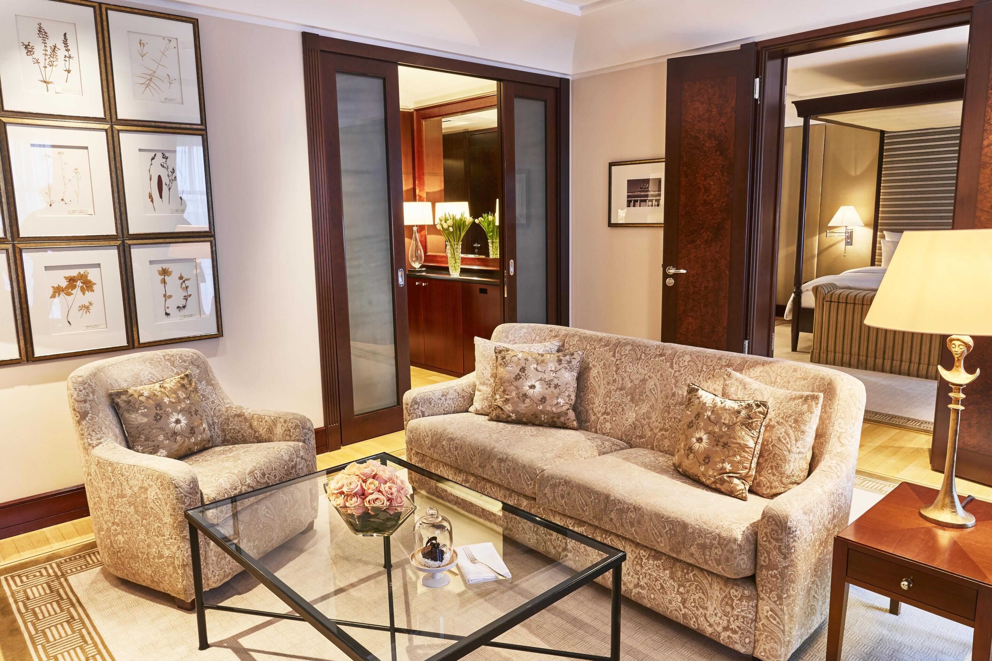 Habitación de hotel con sofá, sillón y una mesa de centro de vidrio con un centro de mesa floral.