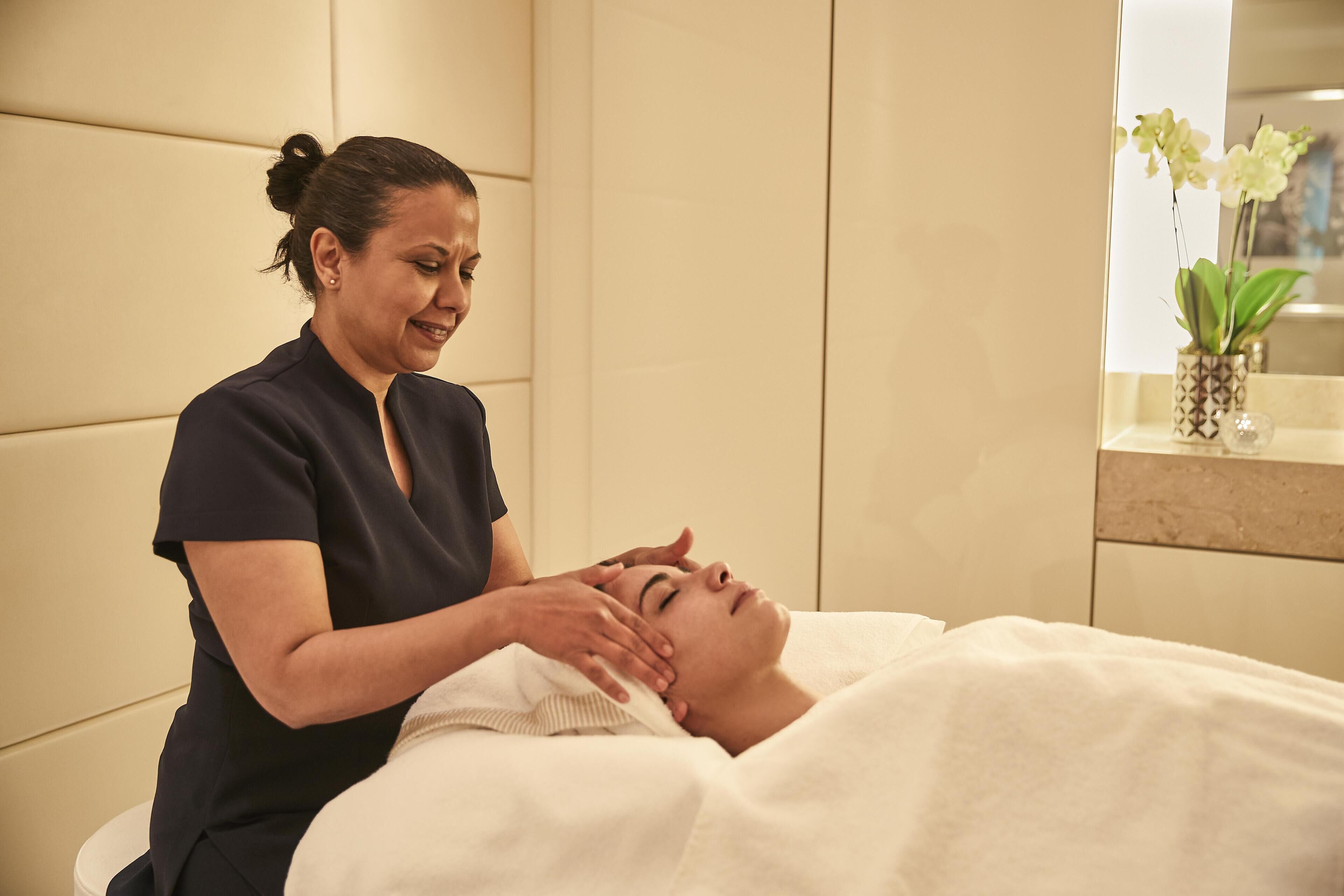 Una persona recibe un tratamiento facial en una habitación con un espejo grande y una planta en maceta.