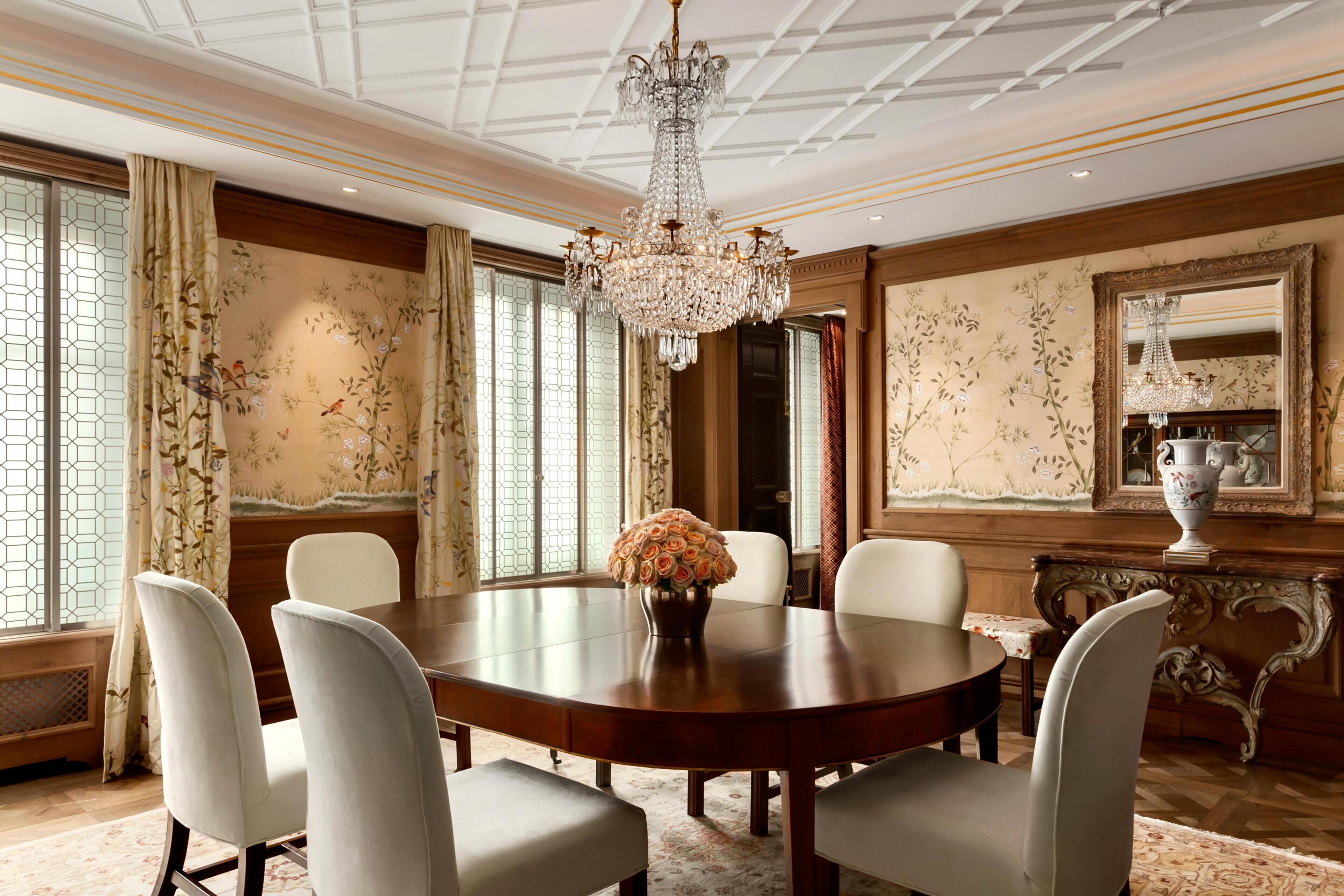 Un comedor formal con una amplia mesa de madera, sillas blancas tapizadas, una araña y papel tapiz con motivos florales.
