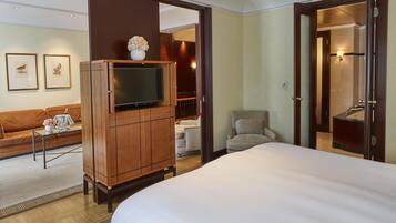 Habitación de hotel con una cama, un televisor sobre un mueble de madera, un sofá, una silla y un baño visible a través de una puerta abierta.