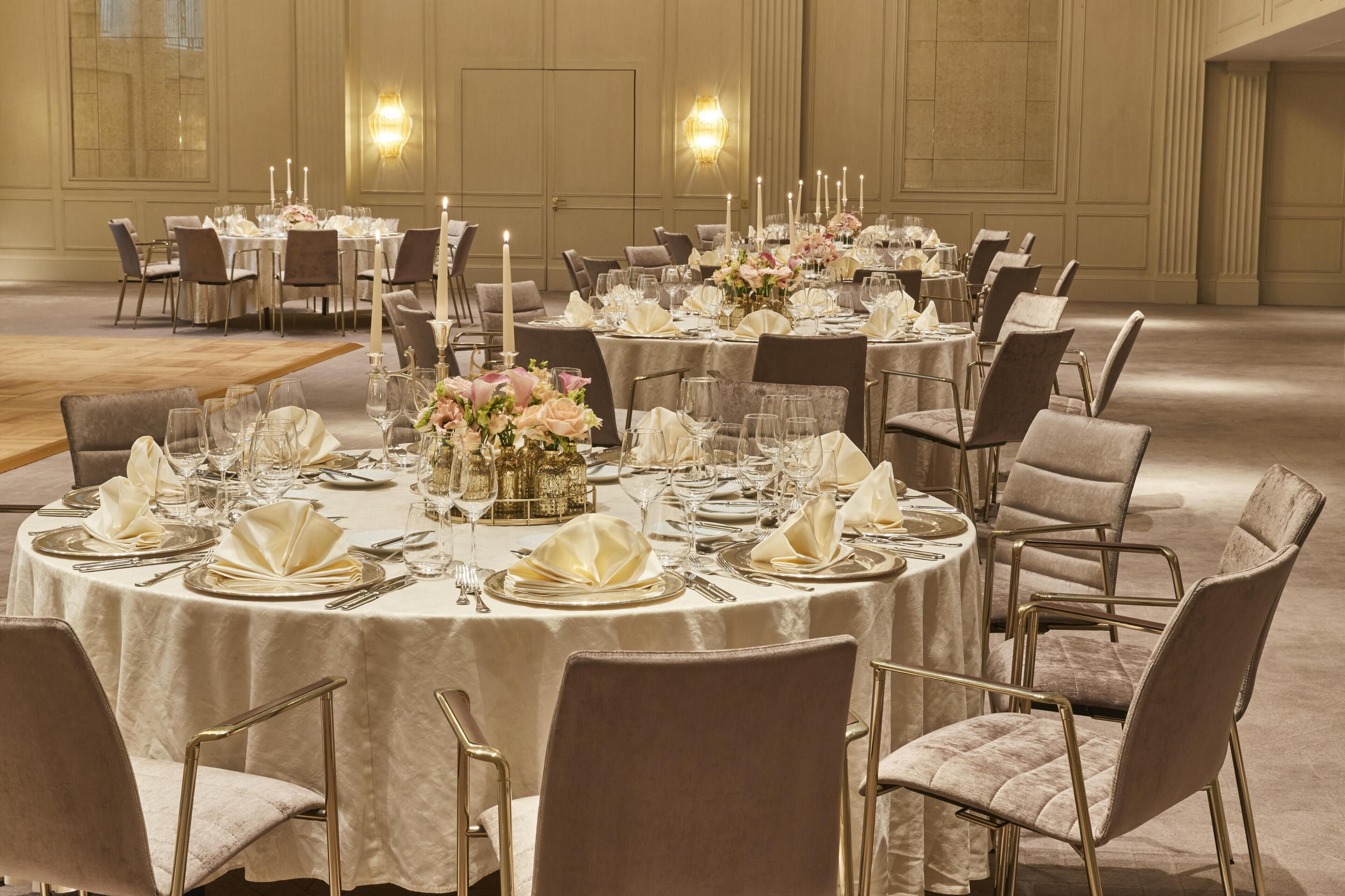 A banquet hall with round tables set for dining, featuring white tablecloths, gold chairs, and floral centerpieces.