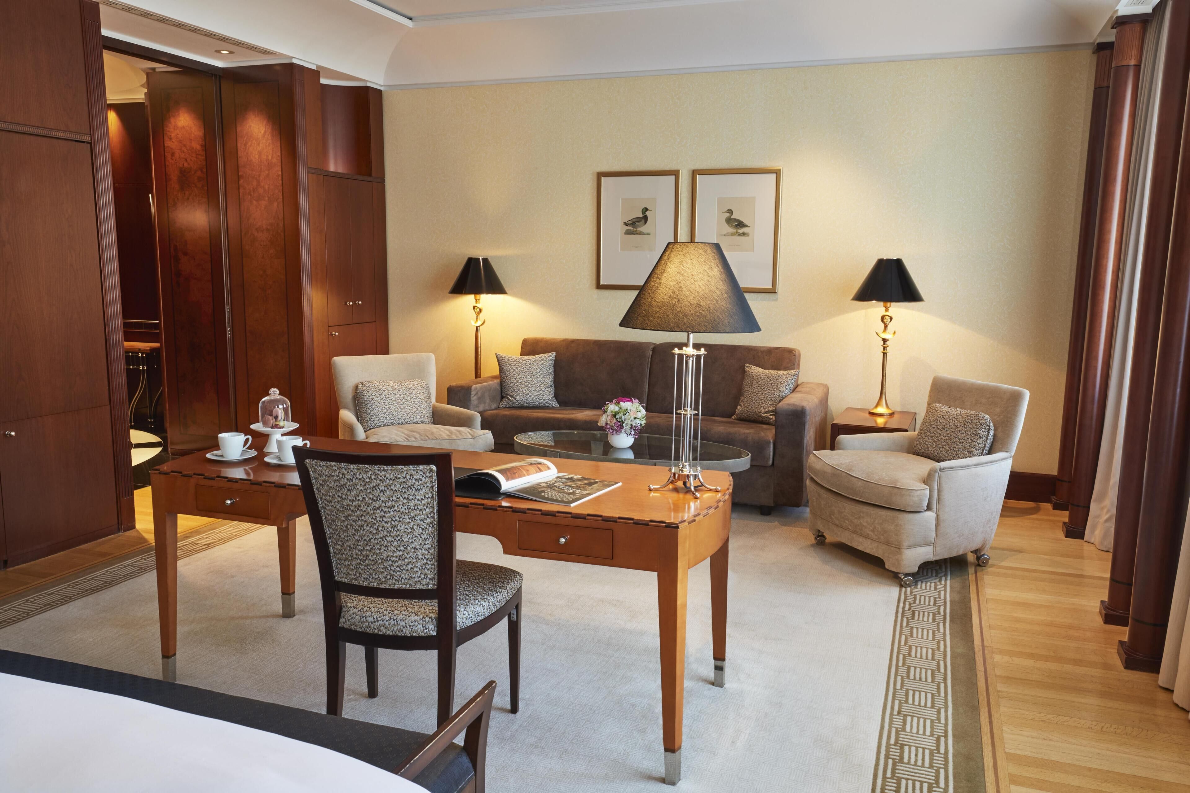 A hotel room with a wooden desk, two armchairs, a sofa, and a coffee table. There are two lamps, a mirror, and framed pictures on the wall.