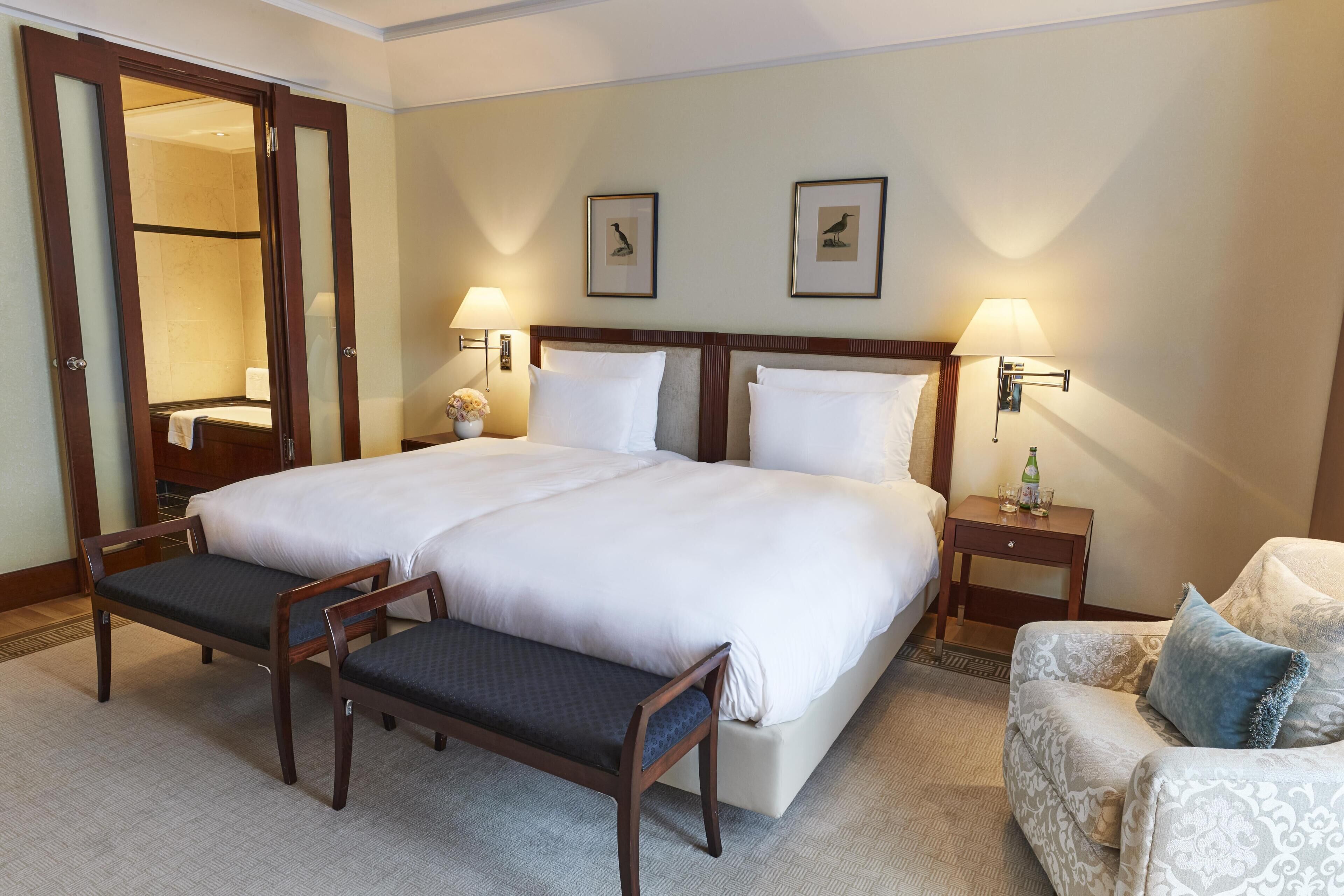A hotel room with a large bed, two bedside tables, a bench, a chair, and two framed pictures on the wall.
