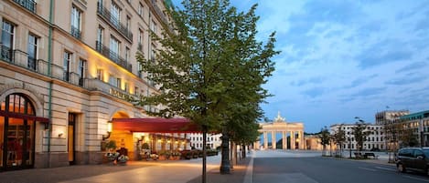 A hotel with a red awning, a tree-lined street, and the Brandenburg Gate in the background.