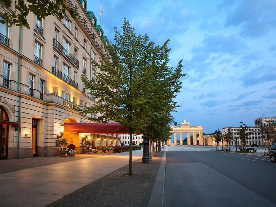 A hotel with a red awning, a tree-lined street, and the Brandenburg Gate in the background.