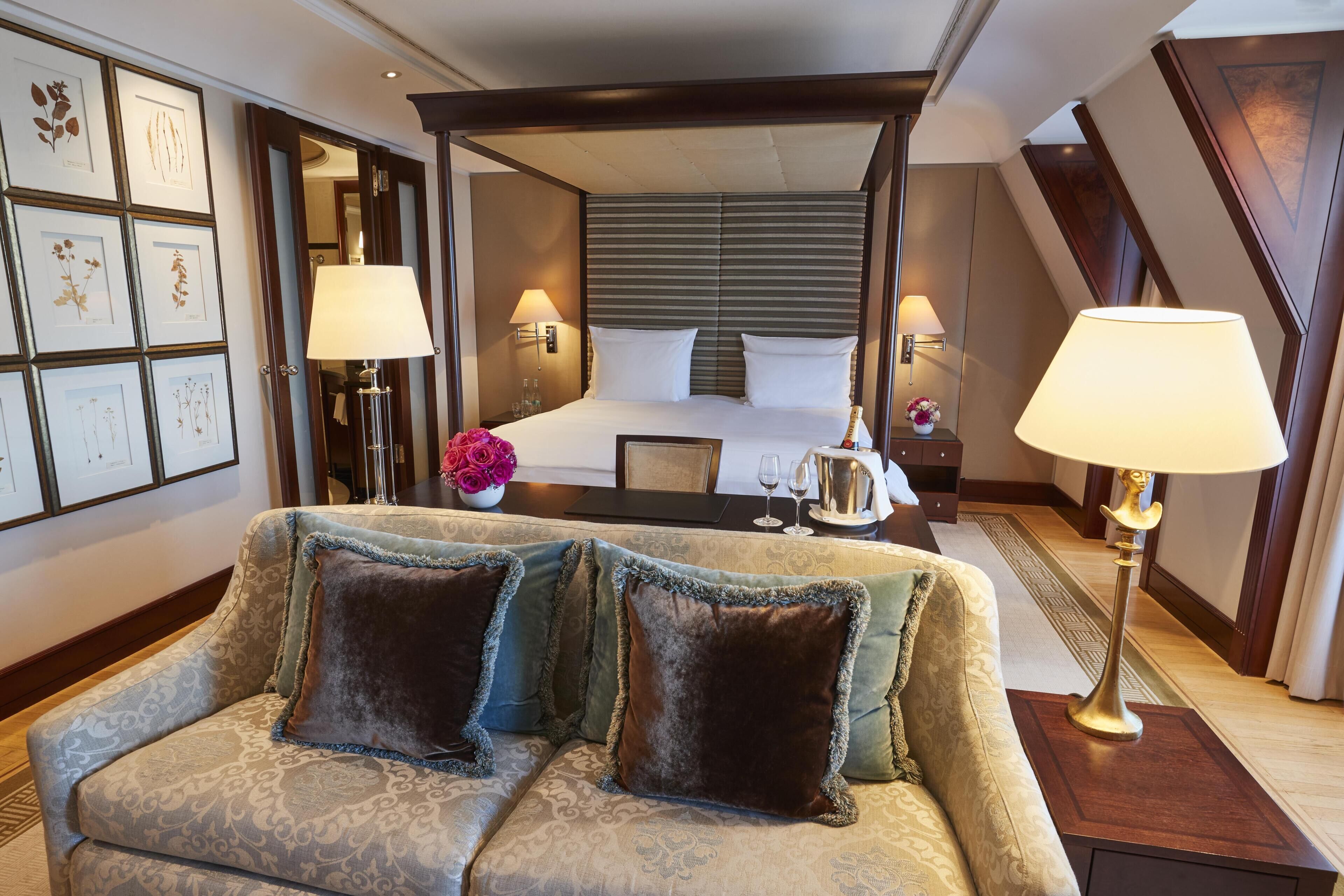 Hotelkamer met een hemelbed, een slaapbank met decoratieve kussens, een nachtkastje met lamp, en ingelijste botanische prints aan de muur.