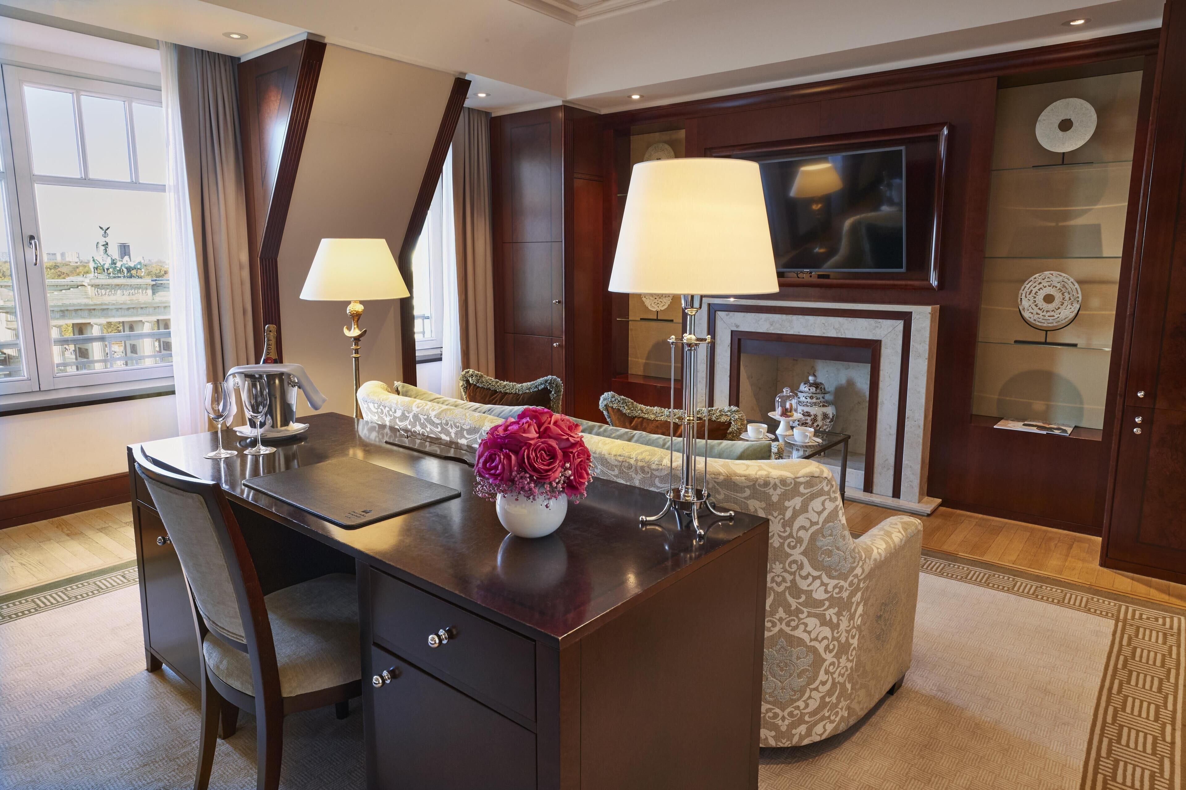 A hotel room with a desk, two lamps, a sofa, a television, a fireplace, and a decorative wall.