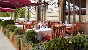 An outdoor dining area with tables and chairs, surrounded by greenery and red umbrellas. - Hotel Adlon Kempinski (Berlin)