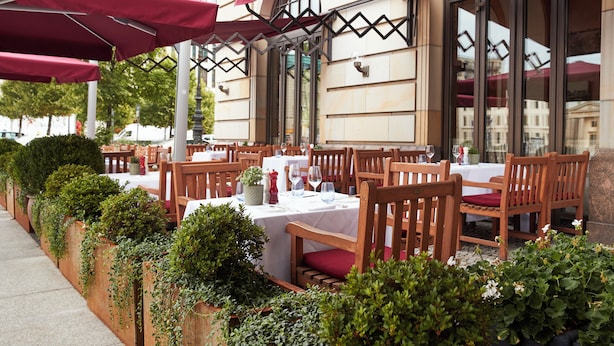 Un espacio de comedor al aire libre con mesas y sillas, rodeado de follaje y sombrillas rojas.