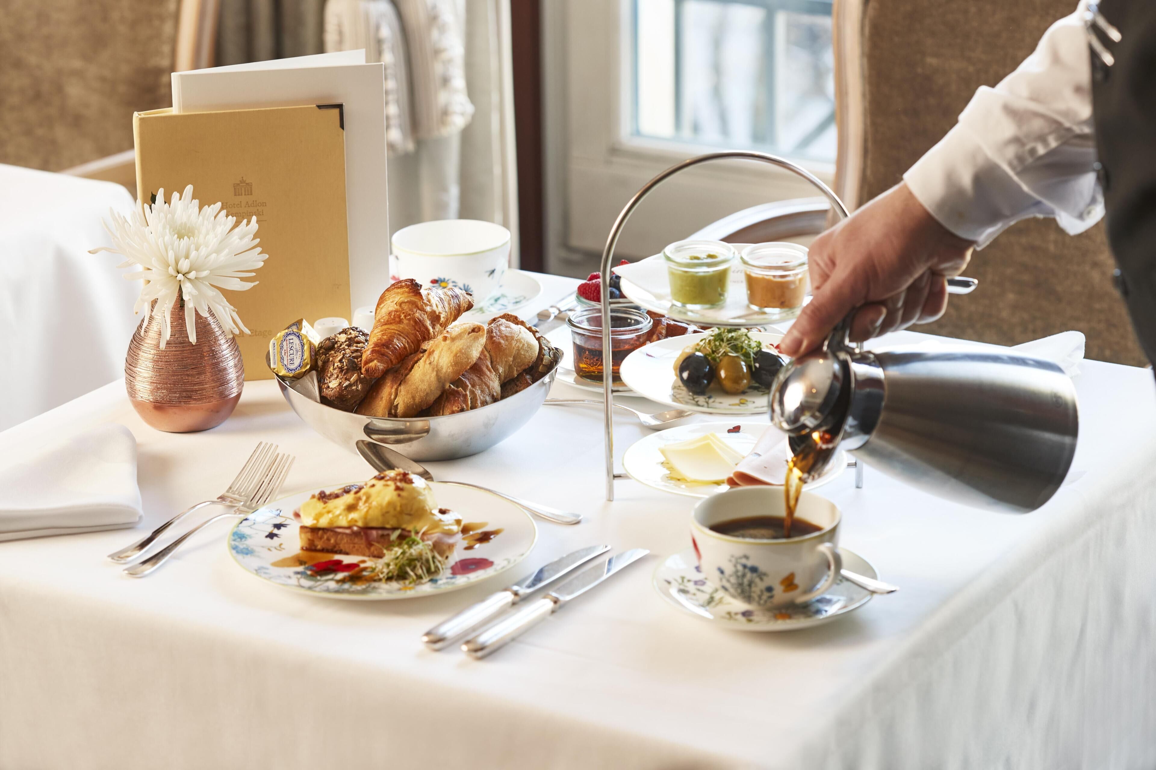 Ein Frühstückstisch mit Croissants, Gebäck, einer Blumenvase und einer Kaffeekanne, aus der Kaffee eingeschenkt wird.