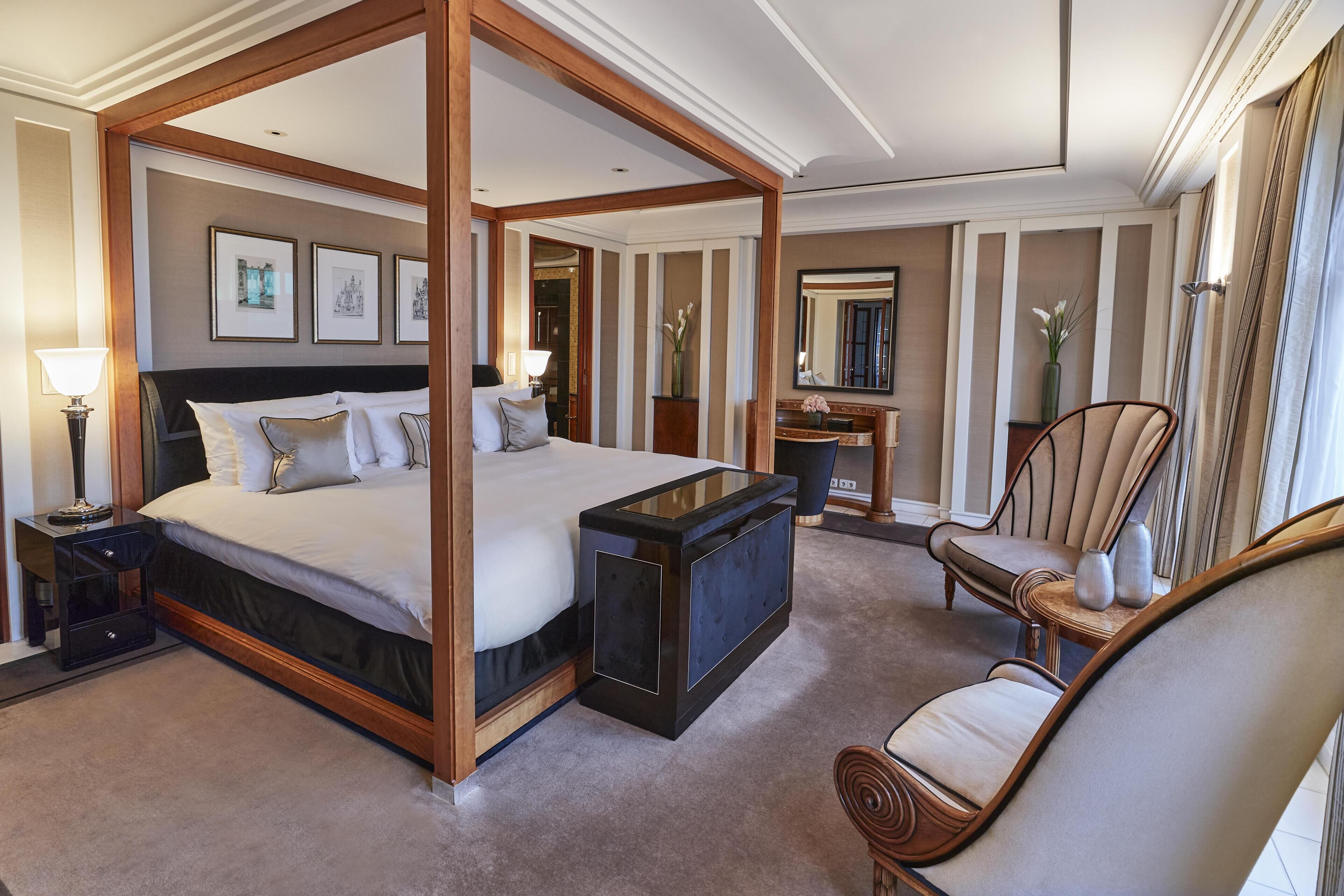 A four-poster bed with a dark headboard, a bedside table with a lamp, a chair with a cushion, and a mirror on the wall.
