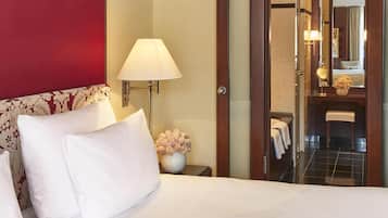 Una camera d'hotel con un letto, un comodino con una lampada, un vaso di fiori e vista sul bagno attraverso una porta aperta.