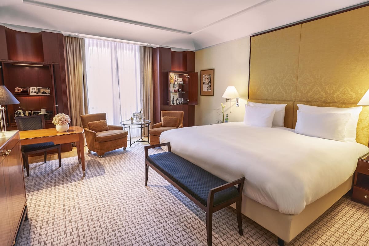 A hotel room with a large bed, a desk with a lamp, a chair, a small table with a vase, and a wardrobe.