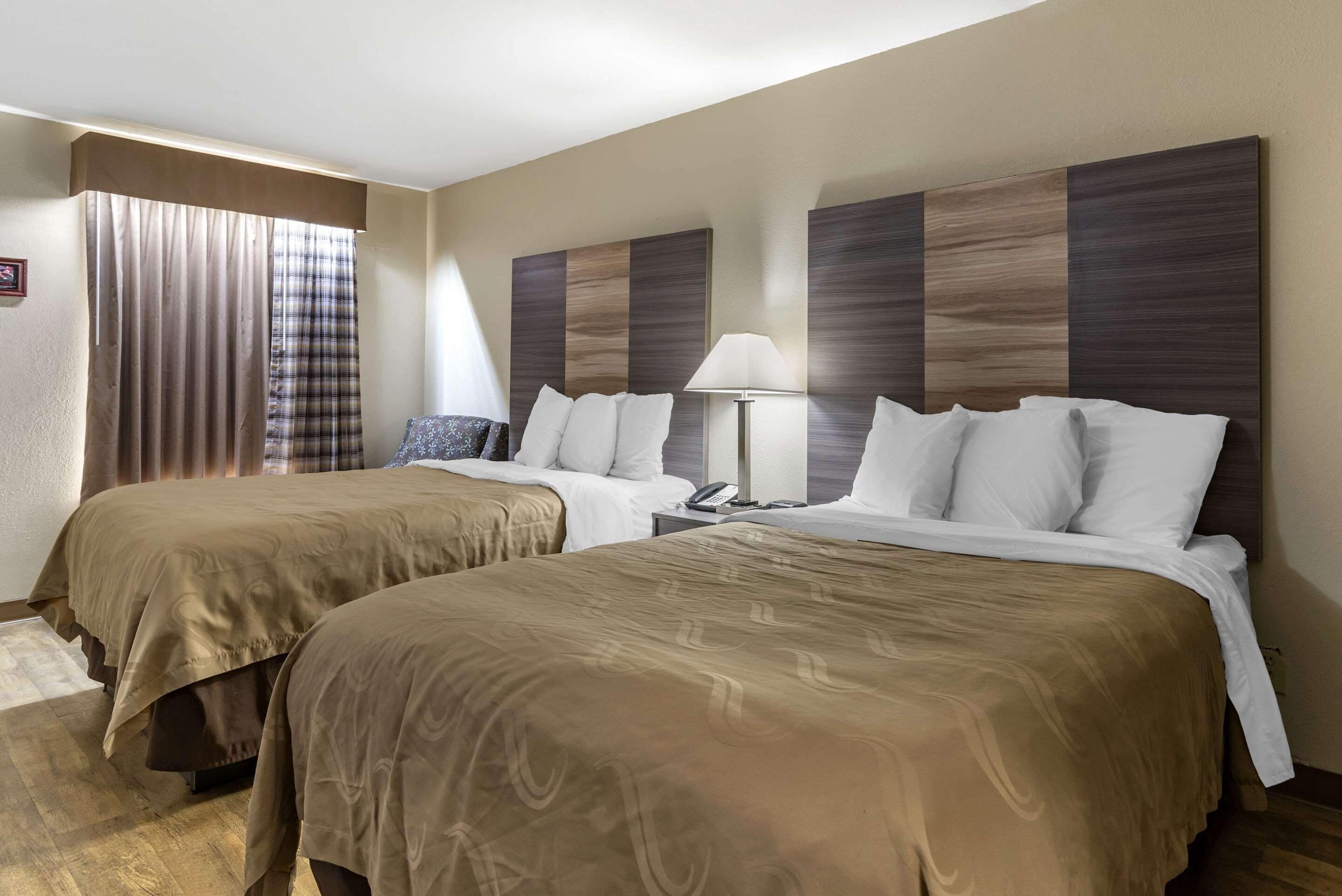 A hotel room with two beds, a nightstand, a lamp, and a window with curtains.