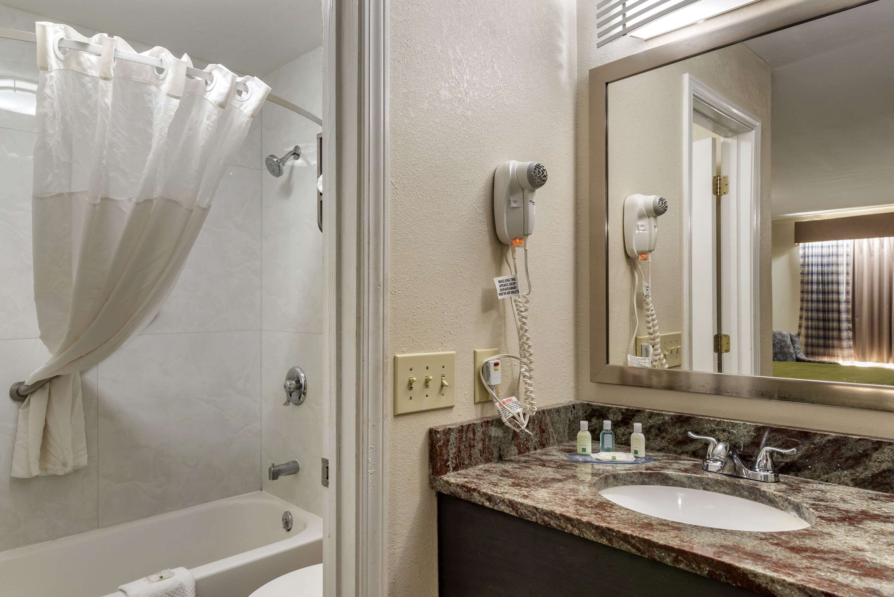 Une salle de bain équipée d’une baignoire, d’un rideau de douche, d’un sèche-cheveux, d’un miroir et d’un lavabo avec des articles de toilette.