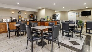 Een restaurantgedeelte van een hotel met meerdere tafels en stoelen, een keuken met houten kasten, en een koelkast.