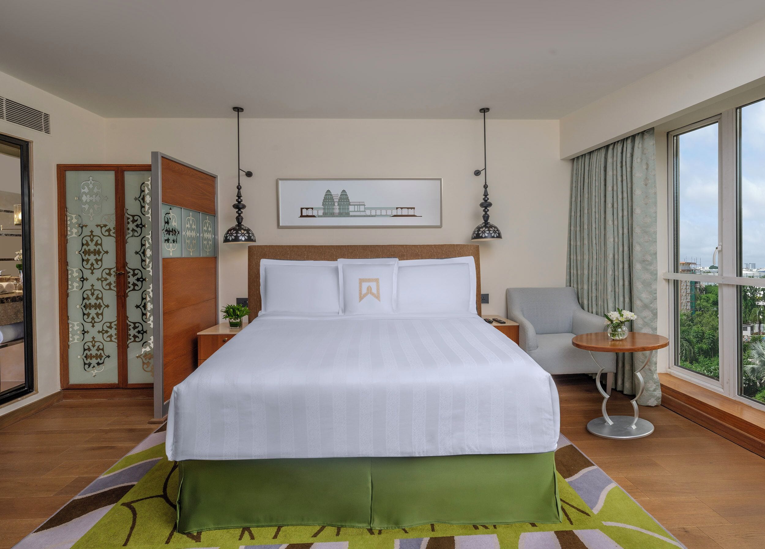Une chambre d’hôtel avec un grand lit, une chaise, une petite table avec un vase, un tableau au mur et une grande fenêtre donnant sur un beau paysage.