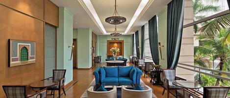 Un vestíbulo de hotel con un sofá azul, dos sillones, una mesa de centro y una araña. Hay mesas y sillas para comer, y se ve una vista de palmeras a través de ventanas grandes.