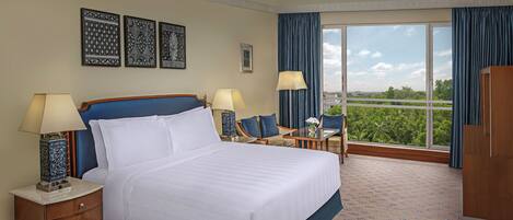 A hotel room with a large bed, two bedside tables with lamps, a small table with chairs, a view of greenery through the window, and decorative wall art.