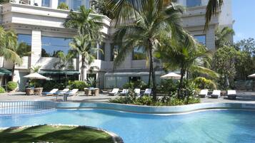 Una piscina con sillones de descanso y palmeras, rodeada por un edificio con varios balcones.