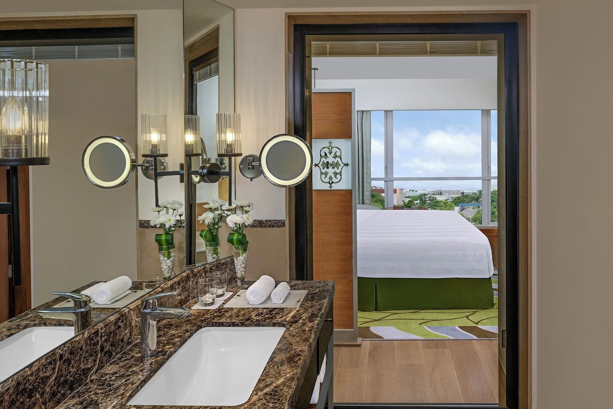 A hotel bathroom with a marble sink, multiple mirrors, and a view of a balcony with a bed.