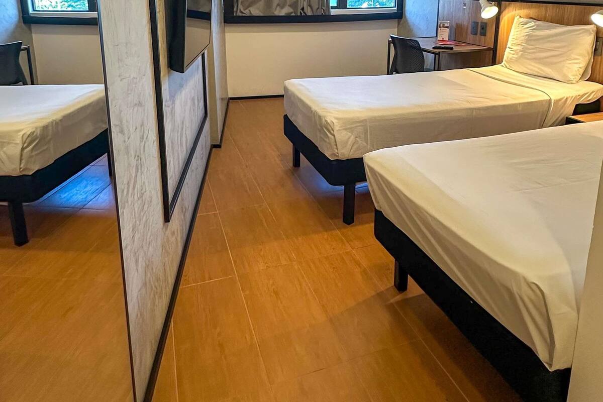 A hotel room with two single beds, a wooden floor, a small desk with a chair, and a window with curtains.